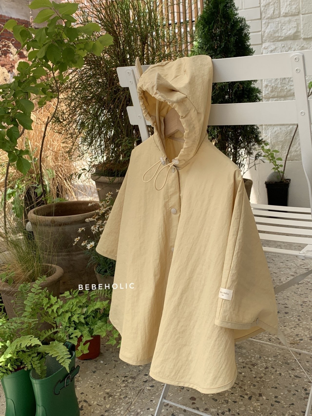 Ein beigefarbener Tierregenmantel mit Kapuze 6 m–24 m – Der perfekte Regenponcho für kleine Abenteurer von Bebe Holic steht auf einem weißen Stuhl in einer Außenumgebung, umgeben von Töpfen mit verschiedenen Pflanzen und Grünpflanzen. Der Poncho hat einen Knopf vorne, eine Kapuze mit Kordelzug und ist aus einem leichten wasserdichten Material gefertigt. Das Wort „BEBEHOLIC“ ist auf dem Stuhl sichtbar.