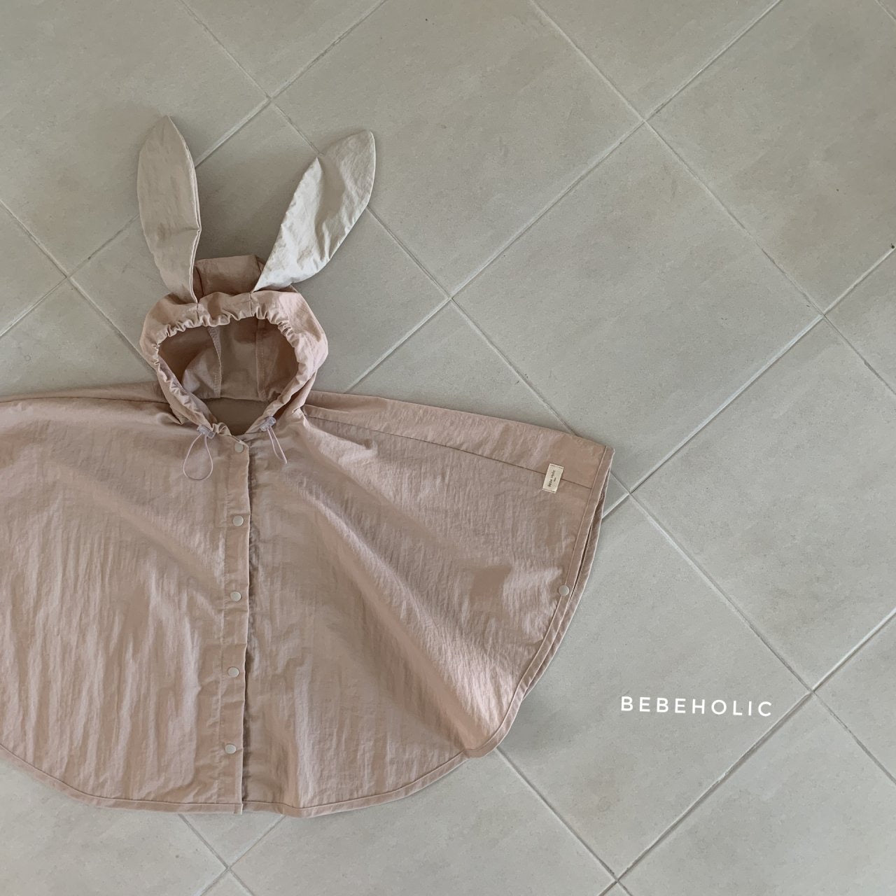 Ein hellrosa Tierregenmantel 6 m–24 m – der perfekte Regenponcho für kleine Abenteurer mit einer Kapuze mit Hasenohren, ausgebreitet auf einem hellgrauen Fliesenboden. Der Poncho hat Knöpfe auf der Vorderseite, ist aus wasserdichtem Material und trägt in der unteren rechten Ecke die Aufschrift „Bebe Holic“.