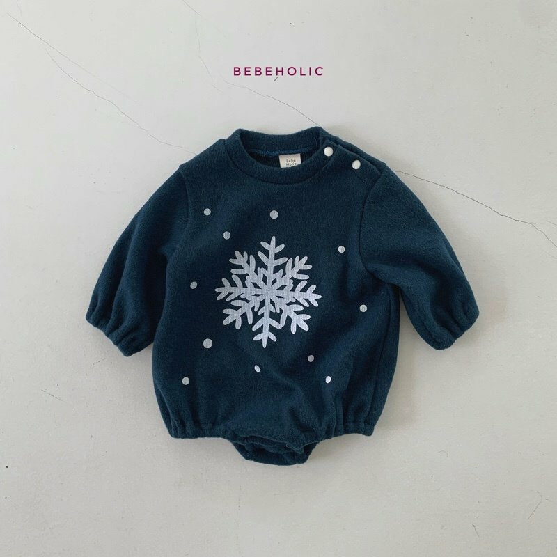 Ein dunkelgrüner Baby-Strampler mit langen Ärmeln, auf dessen Vorderseite eine große weiße Schneeflocke und verstreute weiße Punkte abgebildet sind. Der Markenname „Bebe Holic“ ist in violetter Schrift über dem Strampler aufgedruckt. Der Hintergrund ist eine schlichte, helle Oberfläche mit dezenten Rissen. Dieses Star Bonnet Set passt perfekt zu unserem Star Bonnet Set von Bebe Holic.