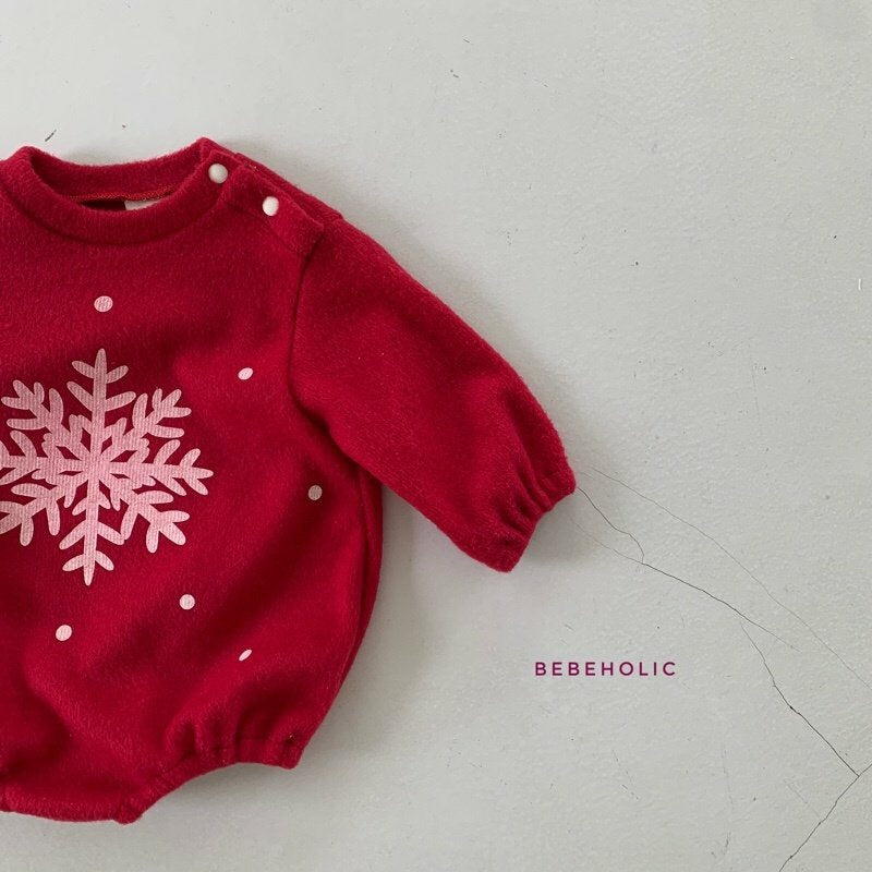 Ein roter Kleinkinderpullover mit langen Ärmeln und einem großen weißen Schneeflockenmuster auf der Vorderseite. Das Star Bonnet Set, Teil der Bebe Holic-Linie, hat Knopfverschlüsse an der linken Schulter und auf der rechten Seite ist „BEBEHOLIC“ in kleinen Großbuchstaben aufgedruckt.
