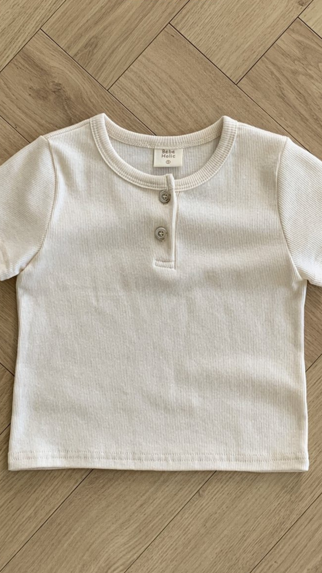 Ein kurzärmliges, cremeweißes geripptes ST-T-Shirt liegt flach auf einem Holzboden mit Fischgrätenmuster. Das ST-T-Shirt hat einen runden Halsausschnitt und zwei Knöpfe auf der Vorderseite, die stilvolle Knopfleisten am Kragen bilden. Ein kleines Etikett im Halsausschnitt zeigt die Marke „Bebe Holic“ und Größeninformationen.