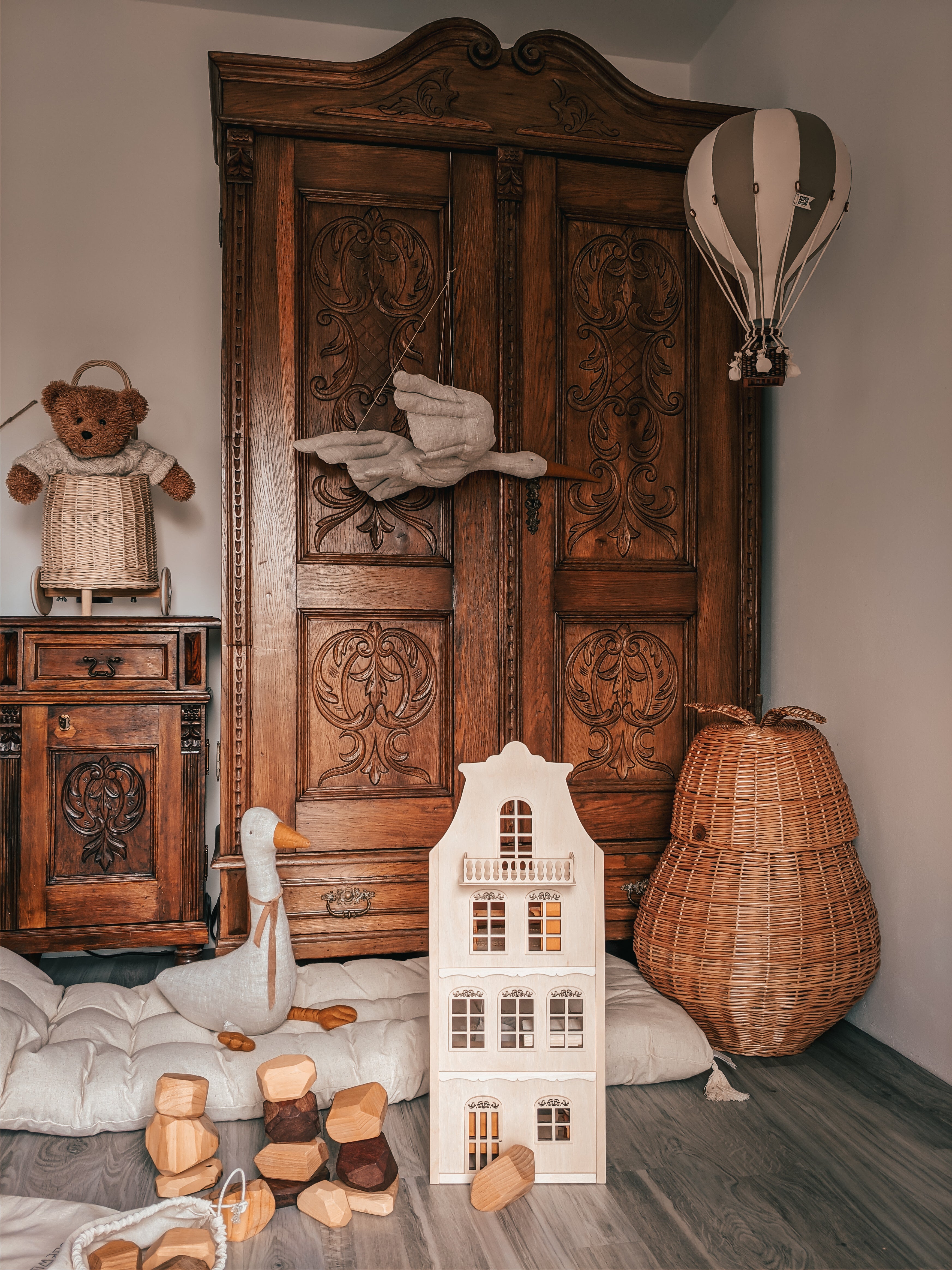 Ein gemütliches Kinderzimmer mit einem aufwendig geschnitzten Holzschrank, einem Weidenkorb und einer Superballoon-Deko-Ballon-Leuchte in BEIGE/DUNKELGRAU. Der Boden ist mit einem großen weißen Puppenhaus, Plüschtieren, darunter einem Teddybären und einem Schwan, und verstreuten Holzklötzen geschmückt.