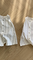 Zwei ärmellose, durchgeknöpfte Bebe Holic Daisy ST Bodys aus leichtem Stoff liegen auf einem Holzboden. Der linke Body hat ein elegantes Streifendesign mit schwarzen vertikalen Streifen auf weißem Hintergrund, während der rechte rosa vertikale Streifen für einen modischeren Look hat.