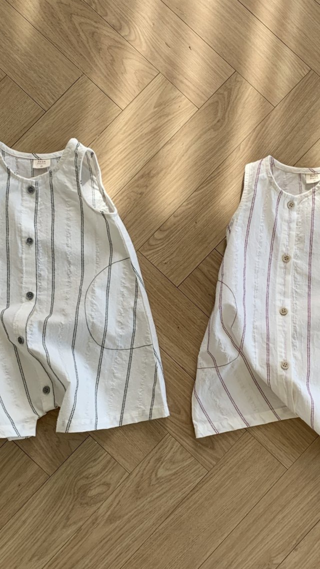 Zwei ärmellose, durchgeknöpfte Bebe Holic Daisy ST Bodys aus leichtem Stoff liegen auf einem Holzboden. Der linke Body hat ein elegantes Streifendesign mit schwarzen vertikalen Streifen auf weißem Hintergrund, während der rechte rosa vertikale Streifen für einen modischeren Look hat.