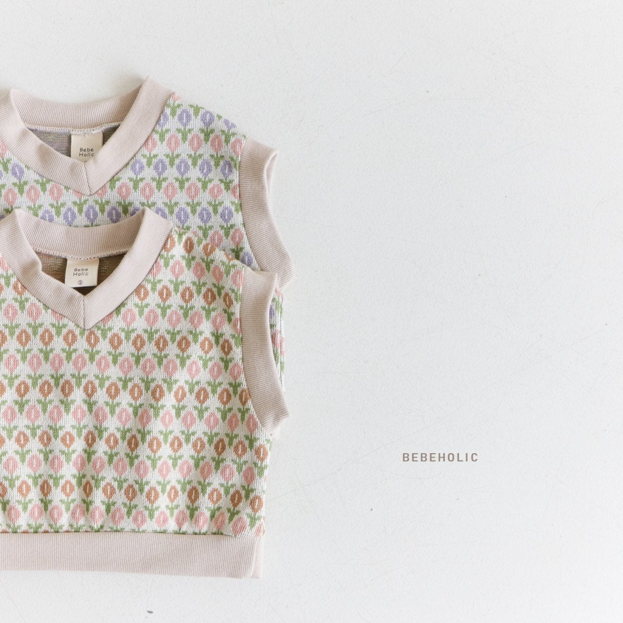Zwei ärmellose Oreo-Strickwesten mit Rauten- und Blumenmuster liegen flach auf einer weißen Oberfläche. Eine Weste weist ein Muster in Hellrosa, Grün und Lavendel auf, während die andere Weste ein ähnliches Design in Zartrosa, Grün und Koralle aufweist. Holen Sie sich mit diesen bezaubernden Westen den Vintage-Look. In der Nähe ist „Bebe Holic“ aufgedruckt.
