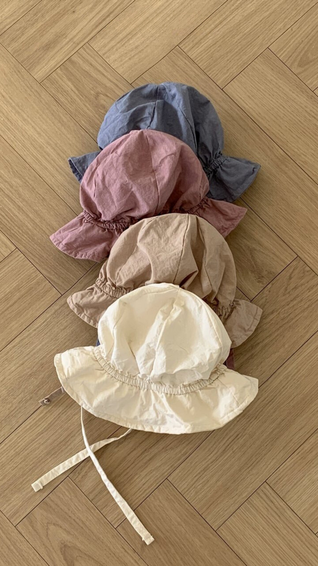 Vier Crunch Bucket Hats in verschiedenen gedeckten Tönen (Blau, Mauve, Beige und Creme) von Bebe Holic sind ordentlich auf einem hellen Holzboden mit Fischgrätenmuster gestapelt, wodurch ihre natürliche Farbpalette hervorgehoben wird.