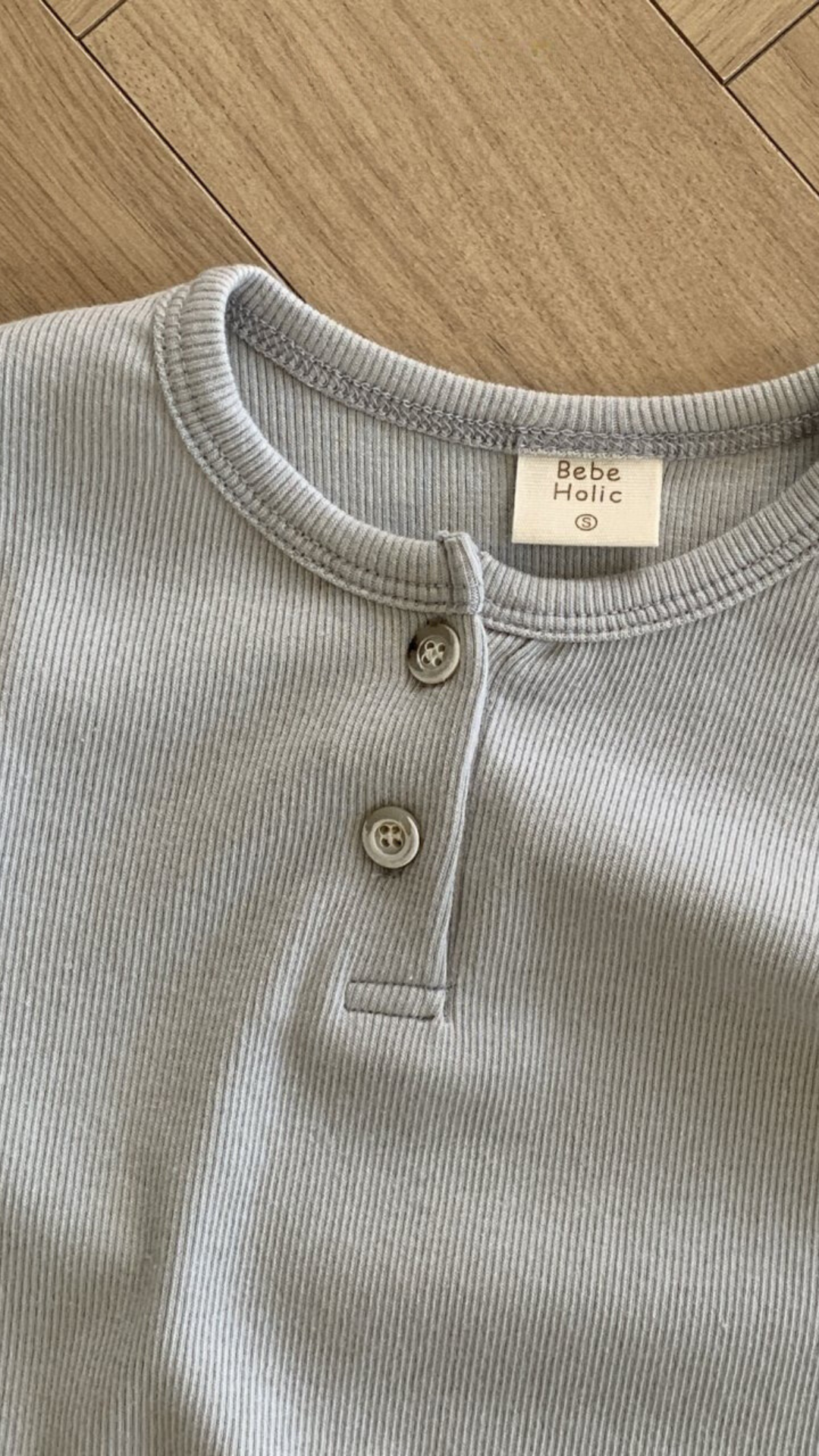Eine Nahaufnahme eines hellgrauen Rippstrickhemds mit zwei Knöpfen zeigt das auf der Innenseite des Kragens aufgenähte Etikett des Bebe Holic ST Tee. Das Hemd aus atmungsaktiver Baumwolle liegt flach auf einer Holzoberfläche mit natürlichem Holzmaserungsmuster.