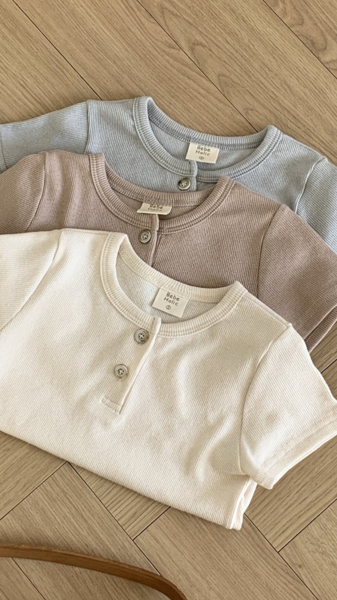 Drei gerippte, langärmelige Bebe Holic ST T-Shirts liegen ordentlich auf einem Holzboden. Die Shirts sind aus atmungsaktiver Baumwolle gefertigt und in den Farben Hellblau, Hellbraun und Creme erhältlich. Jedes hat Knopfleisten am Kragen mit einem oder zwei Knöpfen und ein kleines Etikett mit der Aufschrift „Little Cotton Clothes“.
