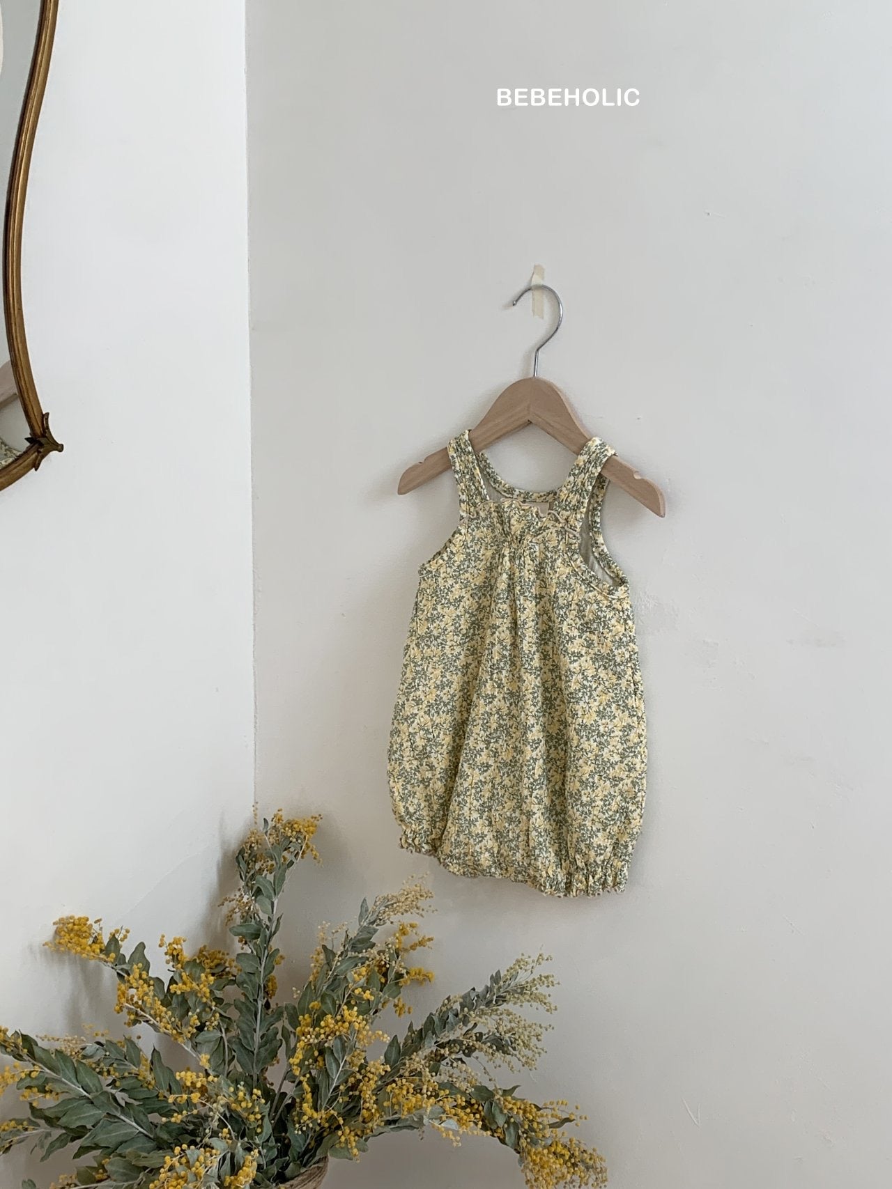 Ein Karen Body von Bebe Holic aus atmungsaktiver Baumwolle hängt auf einem hölzernen Kleiderbügel vor einer hellgrauen Wand. Das verspielte Outfit ist mit kleinen Blumen in Gelb- und Grüntönen geschmückt. Ein kleiner Strauß gelber Blumen liegt darunter und verleiht der Szene einen Hauch von natürlichem Charme.