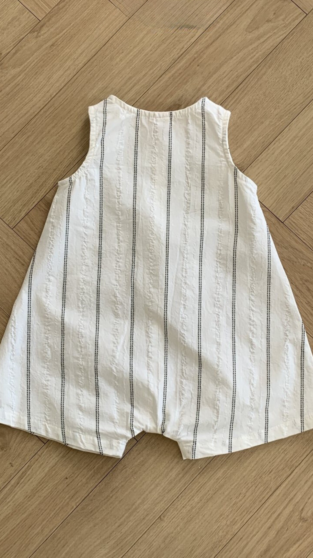 Ein Baby-Strampler liegt flach auf einem Holzboden. Der Daisy ST Body Suit von Bebe Holic ist ärmellos und aus weißem Stoff mit dünnen, vertikalen grauen Streifen. Er hat eine lockere Passform und sorgt für ein bequemes und luftiges Tragegefühl mit einem eleganten Streifendesign, das zu seinem modischen Look beiträgt.