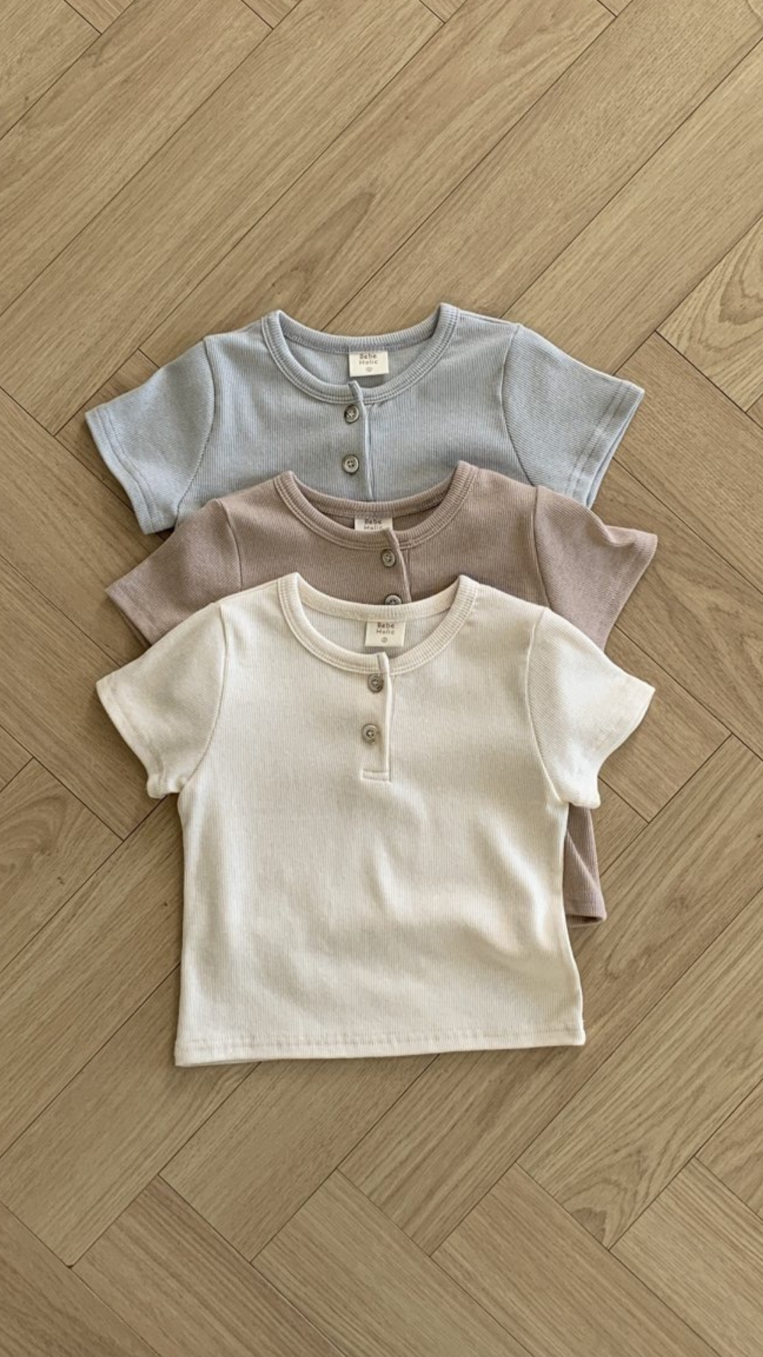 Drei Bebe Holic ST T-Shirts, ordentlich in einem vertikalen Stapel auf einem Holzboden angeordnet. Die Shirts aus atmungsaktiver Baumwolle sind in gedeckten Pastellfarben gehalten: oben hellblau, in der Mitte hellbraun und unten cremefarben. Jedes Shirt verfügt über ein kleines Knopfleisten-am-Kragen-Detail.
