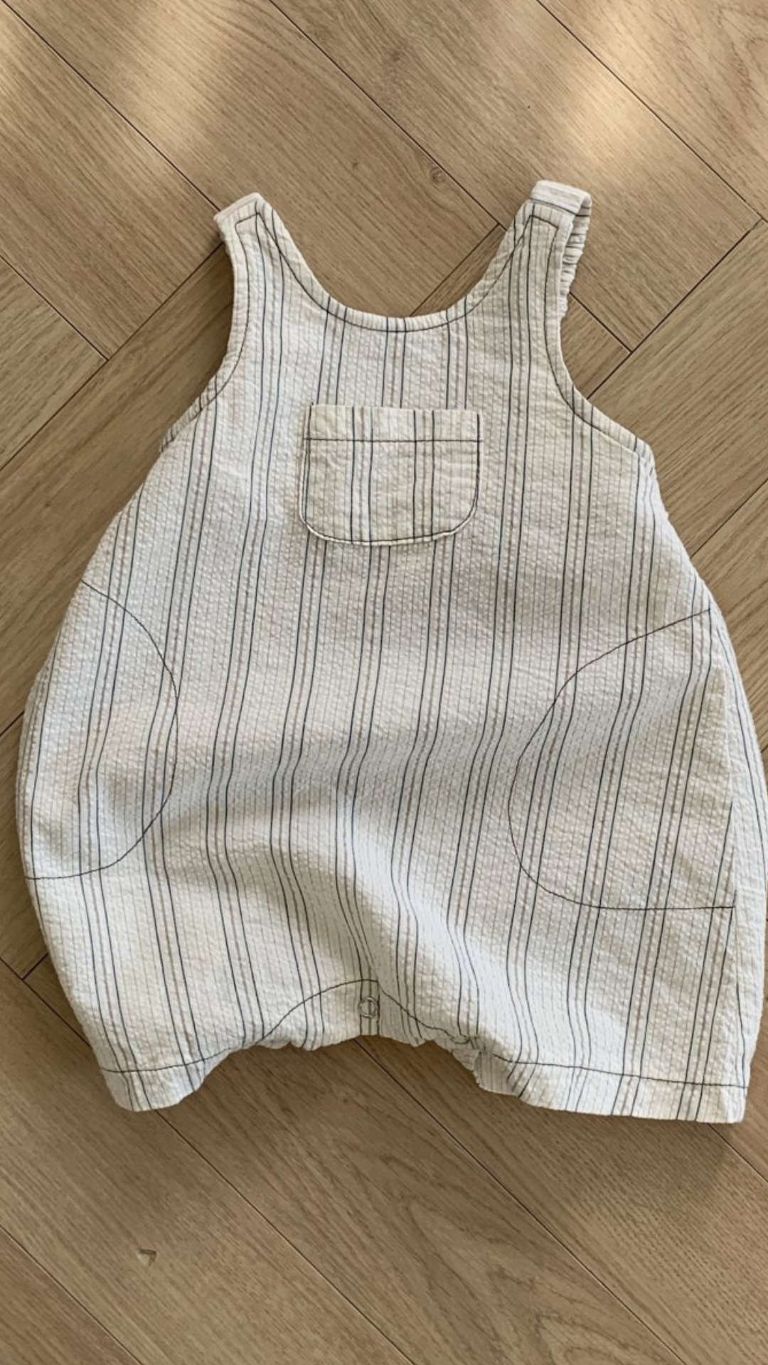 Ein beigefarbener Bebe Holic Ariel St Body mit dünnen vertikalen blauen Streifen, einer kleinen Brusttasche und zwei größeren Seitentaschen liegt flach auf einem Holzboden. Der Strampler hat Schultergurte und elastische Beinöffnungen, perfekt für alle, die Naturtöne lieben.