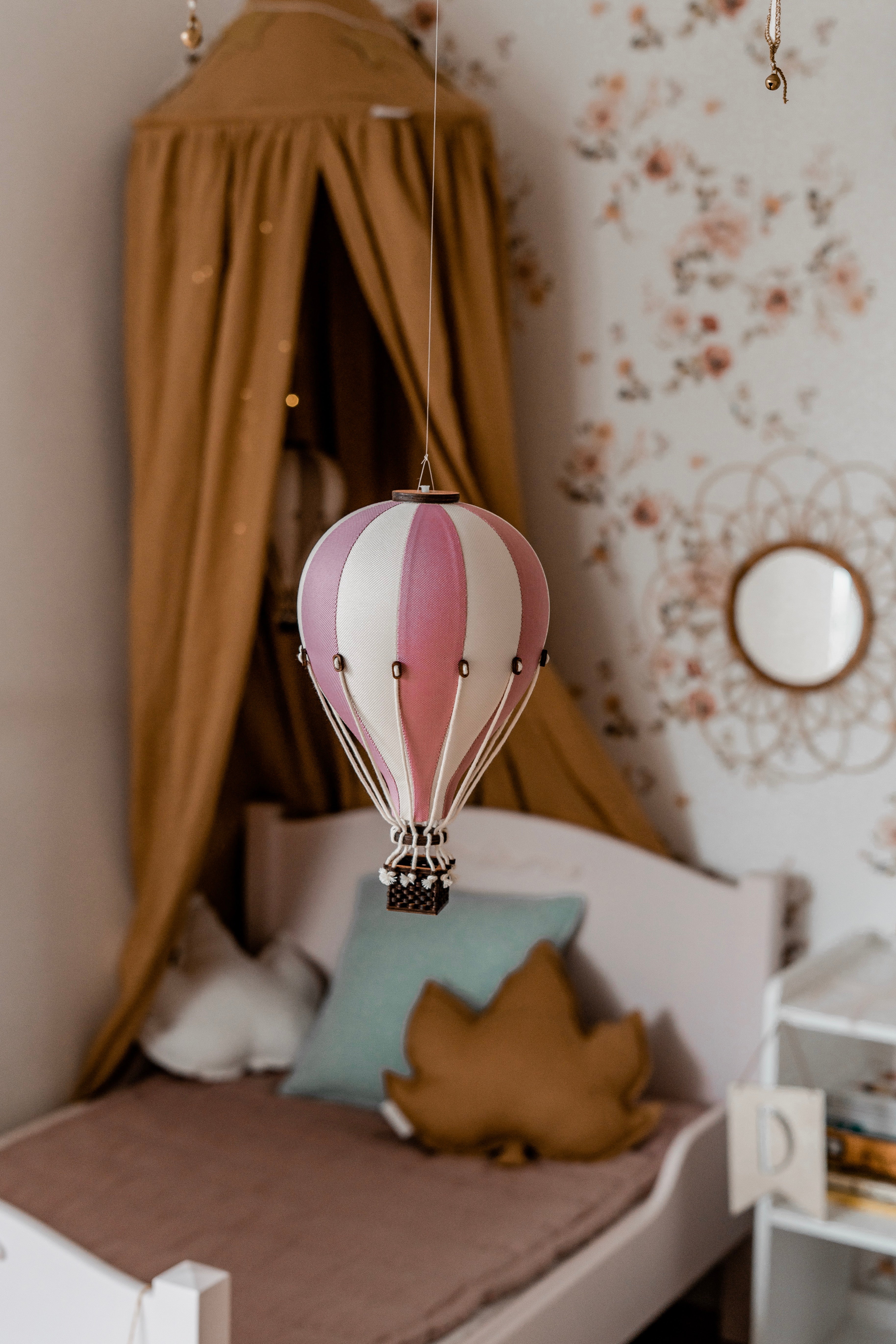 Ein gemütliches Kinderzimmer verfügt über einen hängenden Superballoon-Dekoballon – BEIGE/PUDERROSA. Es gibt ein weißes Bett mit einem weichen senfgelben Baldachin und einer Auswahl an Kissen, darunter ein blattförmiges. Blumentapeten und ein kleiner runder Spiegel verleihen dem Zimmer Charme.