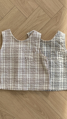 Zwei Summer Check Sleeveless Tees von Bebe Holic aus strukturiertem Stoff mit Gittermuster liegen auf einem Holzboden. Ein Oberteil ist weiß mit rotem Gitter, das andere weiß mit schwarzem Gitter. Jedes Oberteil hat eine kleine Tasche auf der Brust, die einen Hauch modischen Flairs verleiht und perfekt in jede Bebeholic-Kollektion passt.