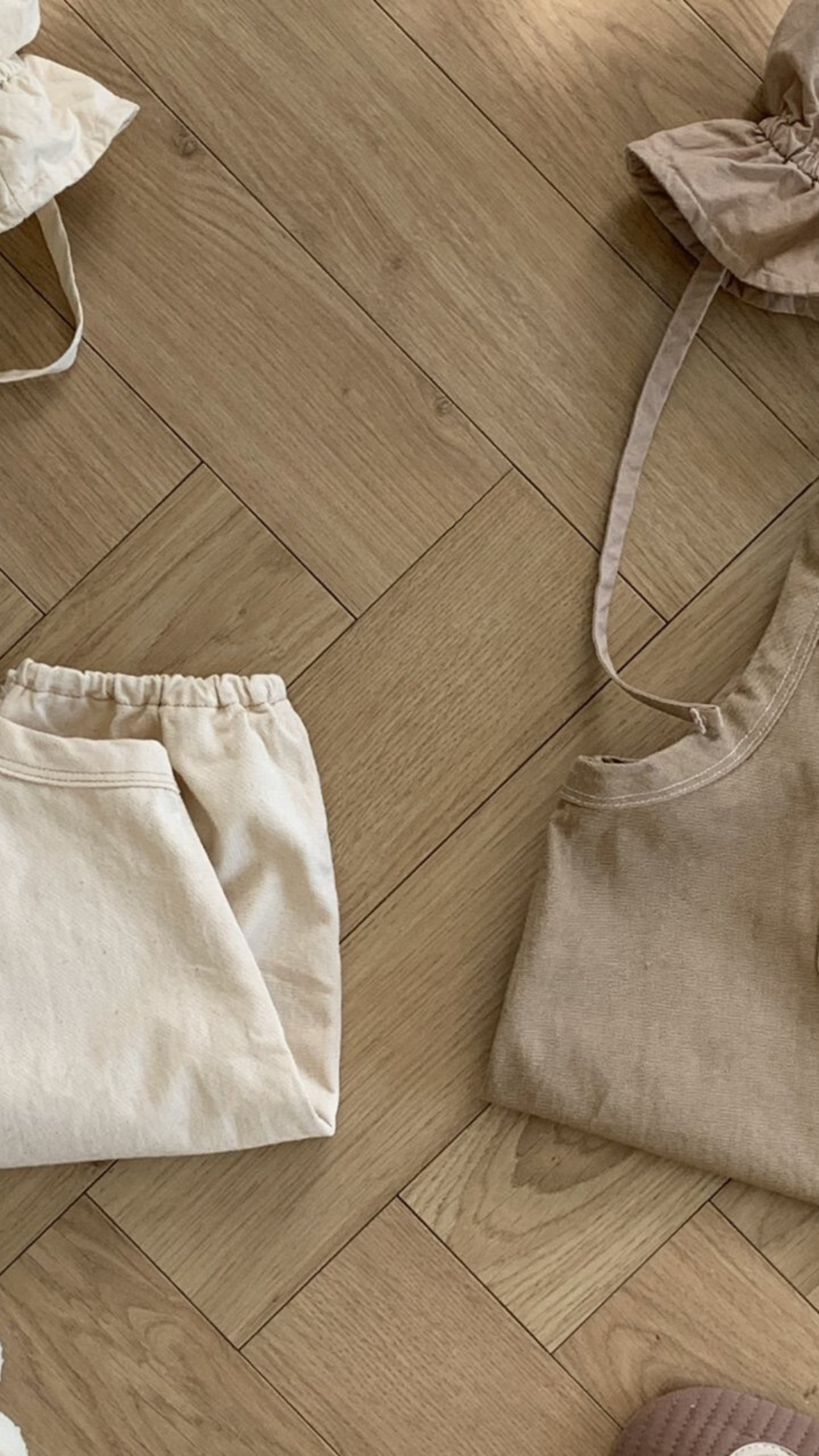 Ein ordentlich gefalteter Twill-Bodysuit mit Taschen von Bebe Holic und ein beiges ärmelloses Oberteil mit verstellbaren Trägern liegen auf einem hellen, hölzernen Boden mit Fischgrätenmuster. Daneben liegen ein Beutel mit Kordelzug und ein gesteppter Artikel, möglicherweise eine Handtasche, alle in ähnlichen neutralen Tönen.